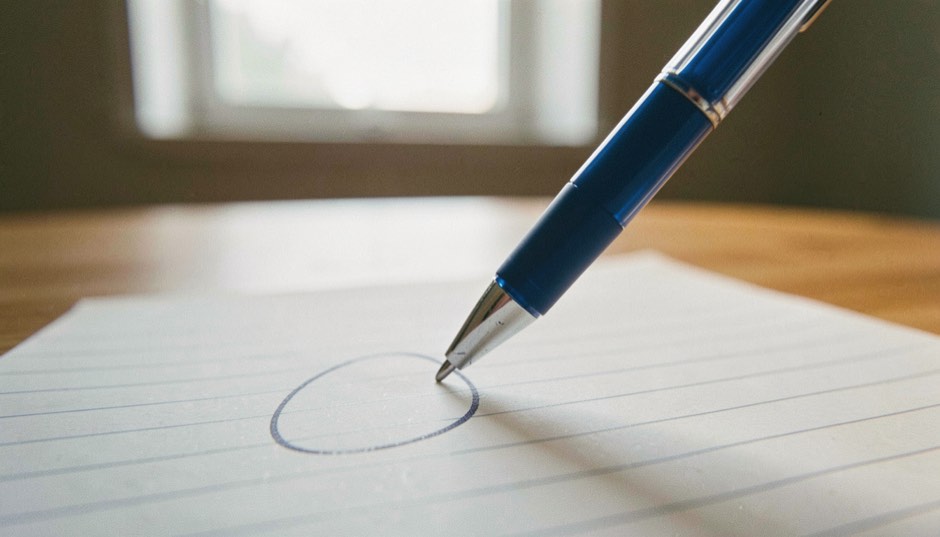Gros plan sur la pointe d’un stylo bleu qui trace une boucle liée sur une page de cahier à lignes, la pointe et la boucle nettes, tout le reste de la page noyé dans le flou, une table de bois clair et la lumière rasante d’une fenêtre derrière.
