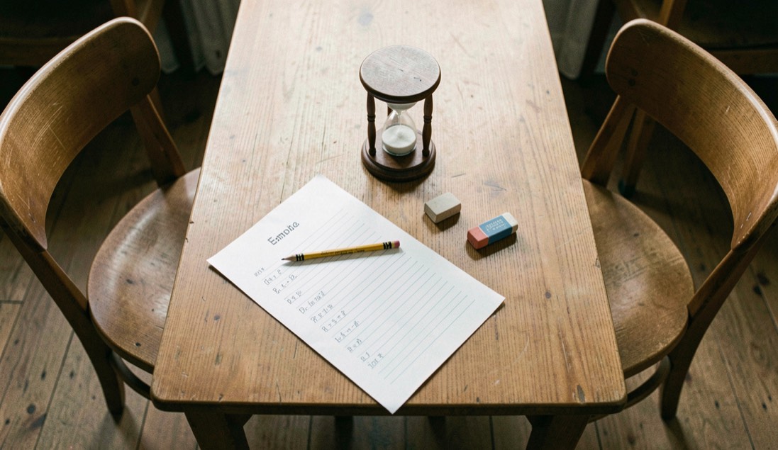 Une table de travail vue de haut, une feuille couverte de lignes d’exercice, un crayon, une gomme et un sablier de bureau posés en triangle, deux chaises vides de part et d’autre.