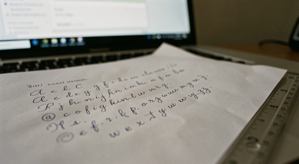 Une feuille couverte d’un alphabet cursive écrit à la main à l’encre, posée devant un clavier d’ordinateur, l’écran flou en arrière plan, une règle graduée le long du bord droit de la feuille.